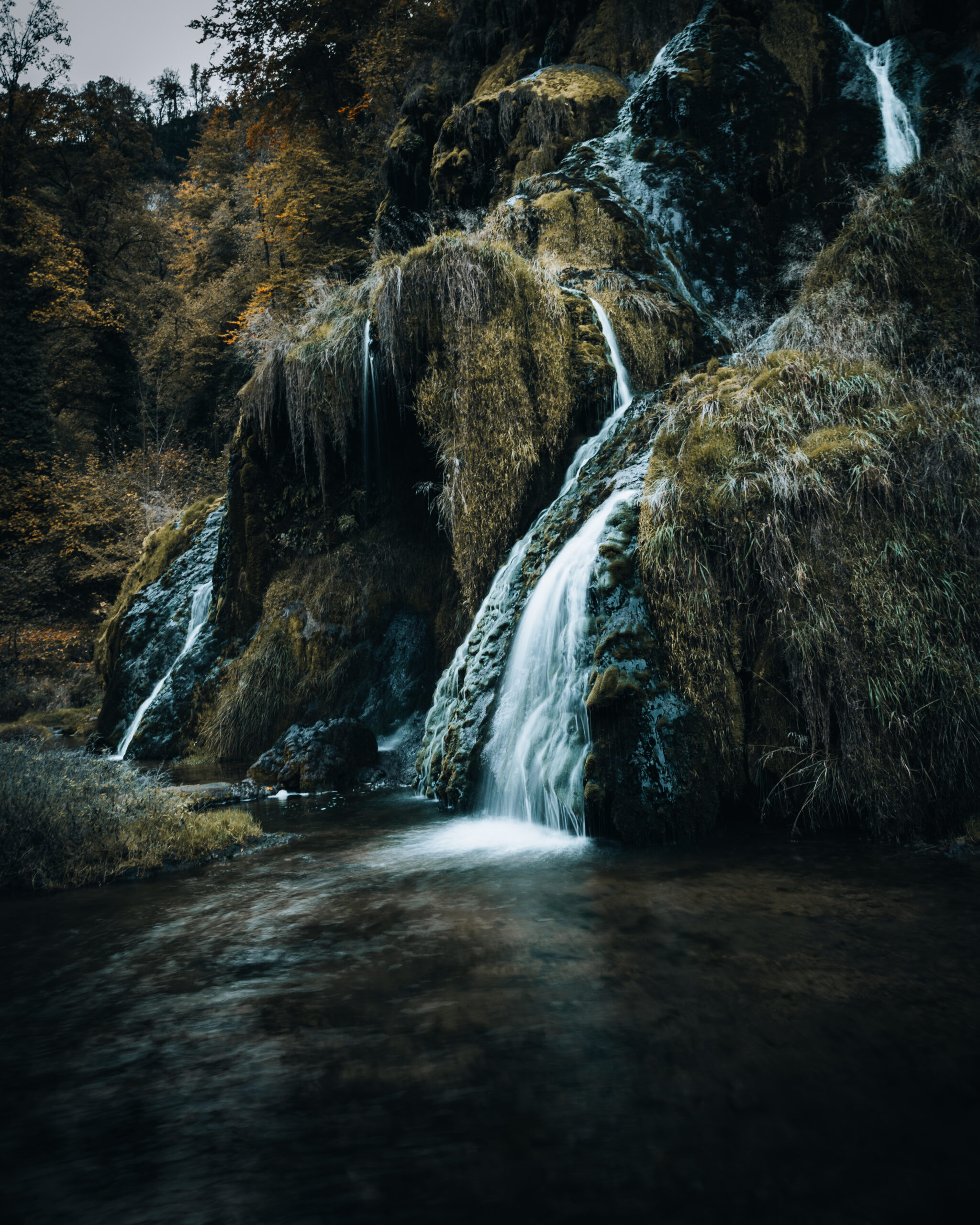 Hidden in the shadowed folds of the forest, water slips quietly over moss and stone, carving time into silence. A place where movement is gentle, light is restrained, and nature speaks only in murmurs.