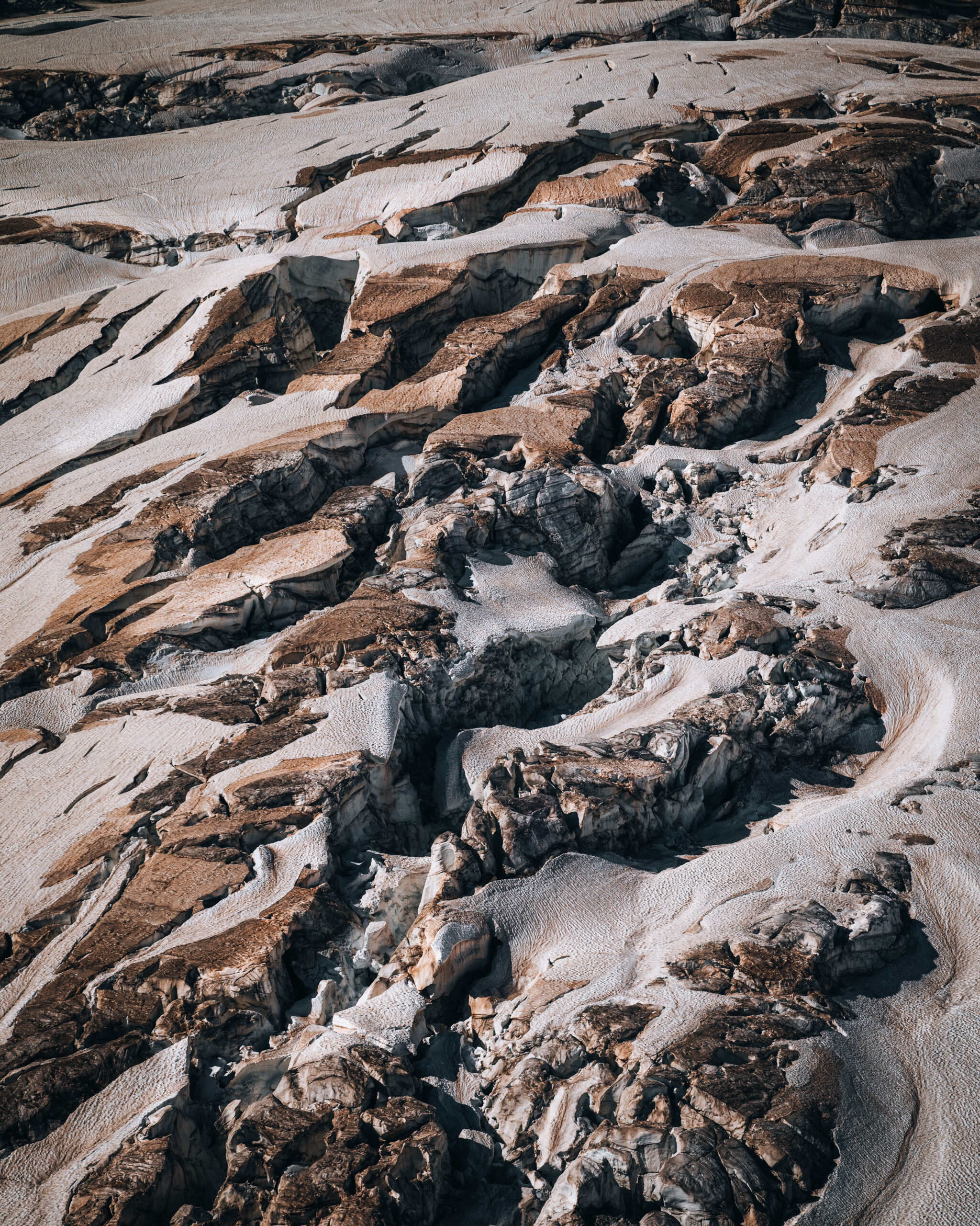 Aerial view of a fractured glacier surface, where exposed rock and melting ice reveal intricate textures and deep crevasses formed by the slow movement of the glacier.